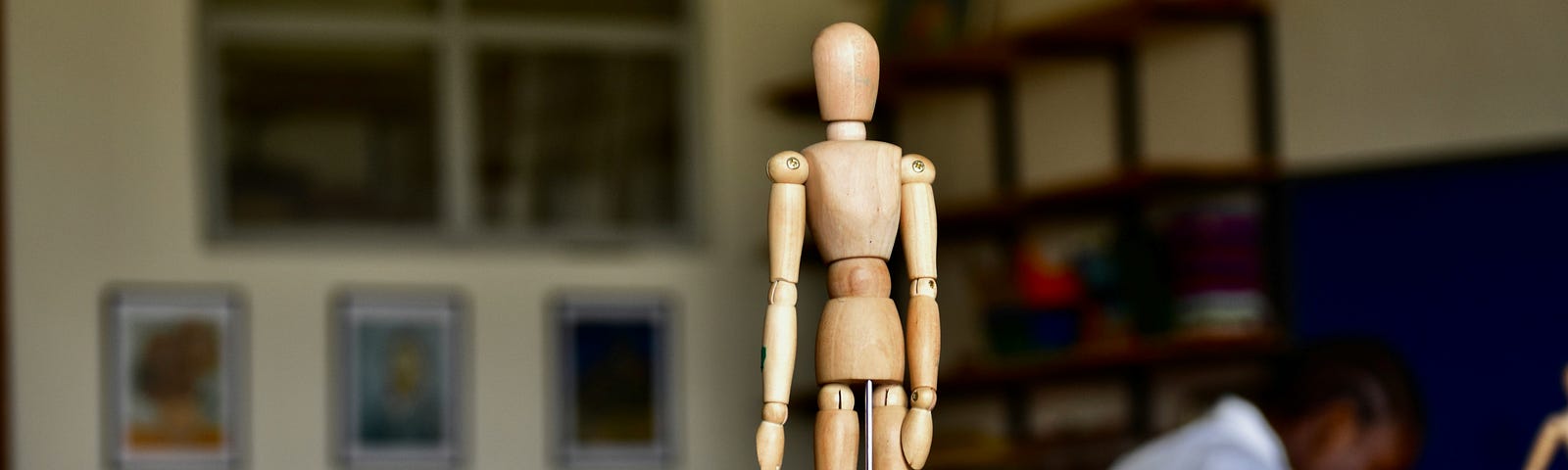 A small wooden figurine of a human, such as those used as drawing or animation references. It sits on a desk. A person can be seen in the background, bent over their work.