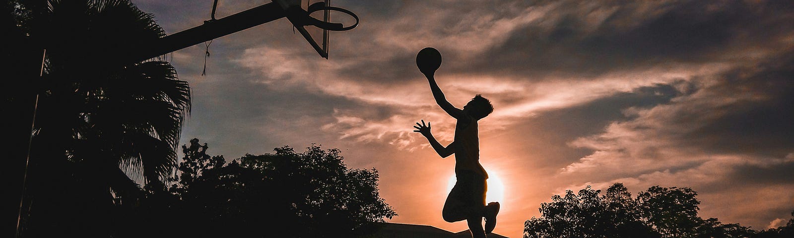 Silhouette of a man shooting a basketball layup. Sunrise behind him.