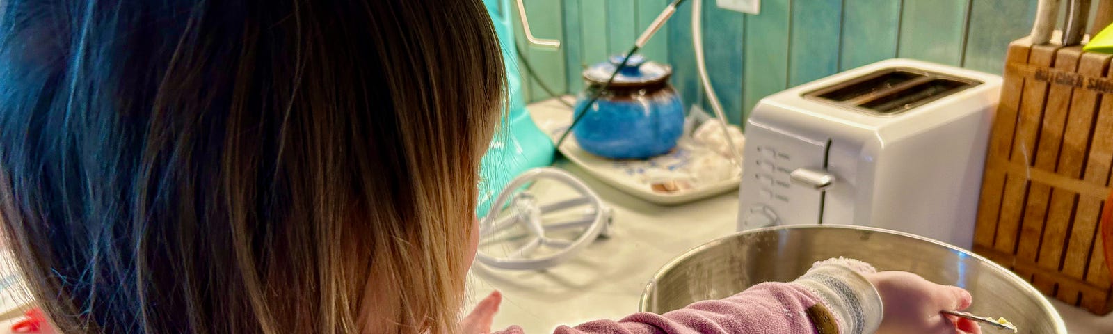 A toddler stands at the counter. She has a mixing bowl in front of her and she is stirring the contents. Her back is to the camera