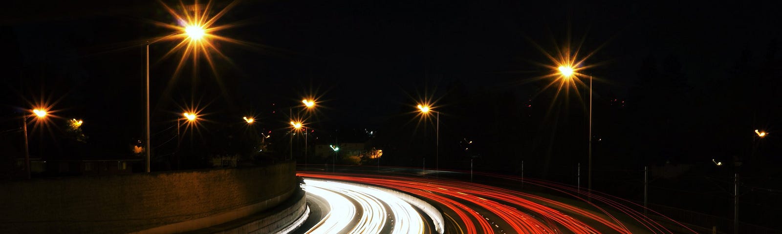 Long exposure image of a highway at night featuring red and white streaks of headlights merging into single rays of light above street lamps set against the night sky.