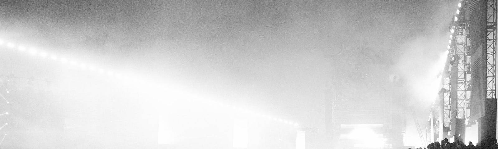 Black & white photo of a large crowd with thick fog in the dark background. Thousands of people are crowded close together, some hold their phones above their heads. On the right of the photo is a screen or thin structure, much taller than the crowd. It’s topped with lights. The photo is busy and full, but dark and mysterious.