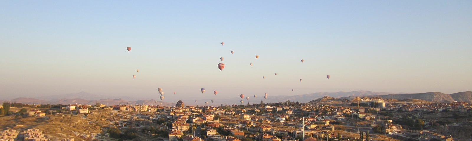 Many hot air balloons flying low over a city at sunrise