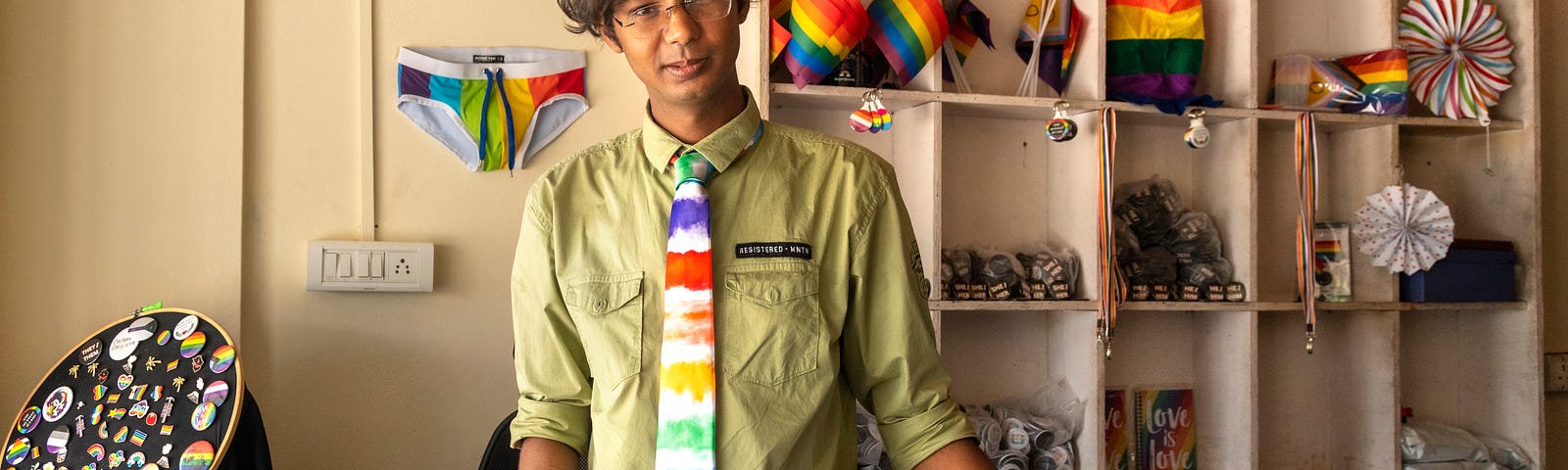 A man wearing a rainbow tie stands behind a store desk. Behind him the walls are covered in rainbow merchandise.