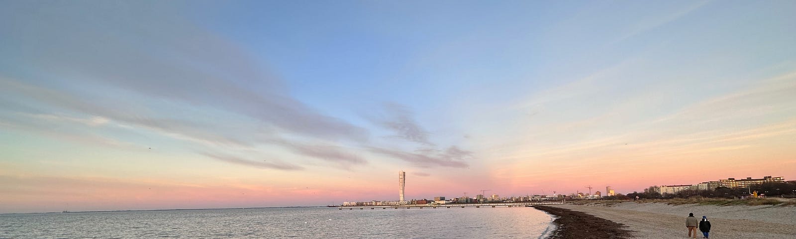 Sunset over Ribersborg beach in Malmö. Turning Torso in the sunset. Two people are walking on the beach. Sea weed washed up on the shores on the beach. Blue skies with some grey white clouds.