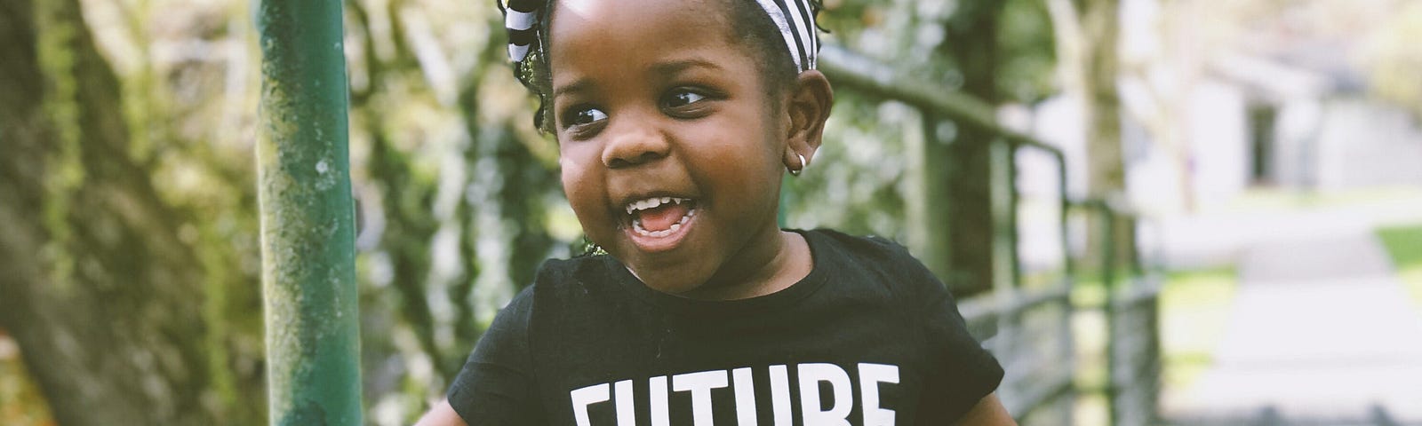 Excited adorable kid, with brown skin and black braids in a striped headband, in a “Future Leader” shirt, on a little bridge