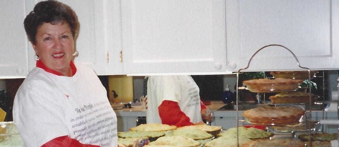 My adoptive mother, wearing a white short-sleeved T-shirt over a red long-sleeved one, smiling and standing by a counter with multiple mincemeat pies on it.