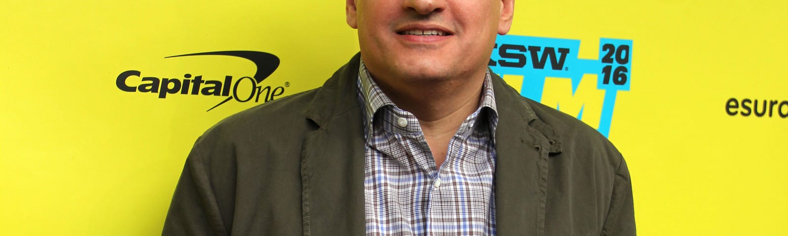 Ted Sarandos standing in front of a primarily yellow step and repeat at SXSW in 2016, he is wearing a brown/greenish jacket, a plaid shirt and blue jeans.