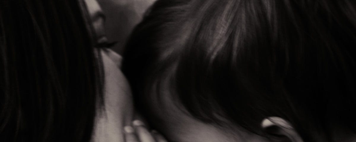 A black and white close up photo of a woman kissing a young child on the forehead while the child’s hand rests on the woman’s cheek