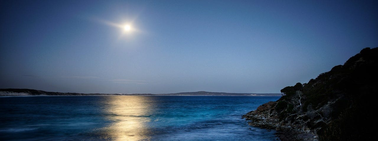 The beautiful blue ocean, calmly lapping at the tocks, as the moon rises, casting her hlow across the waters.