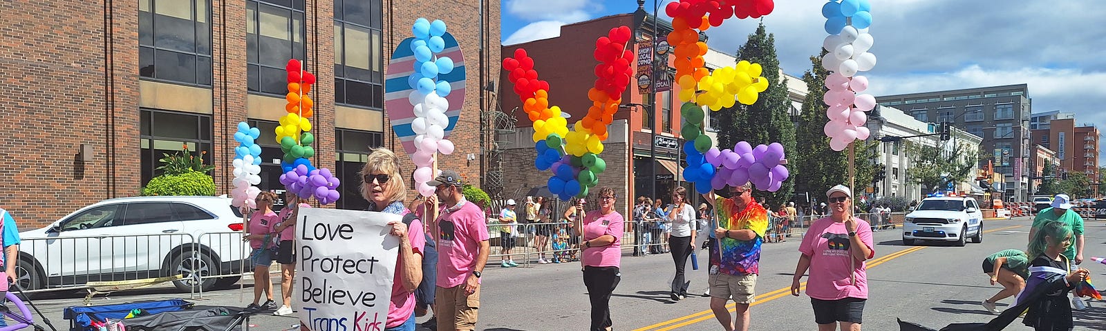 A Pride Parade with people holding balloons that spell out “LOVE” and a woman holding a sign that reads, “Love Protect Believe Trans Kids.”