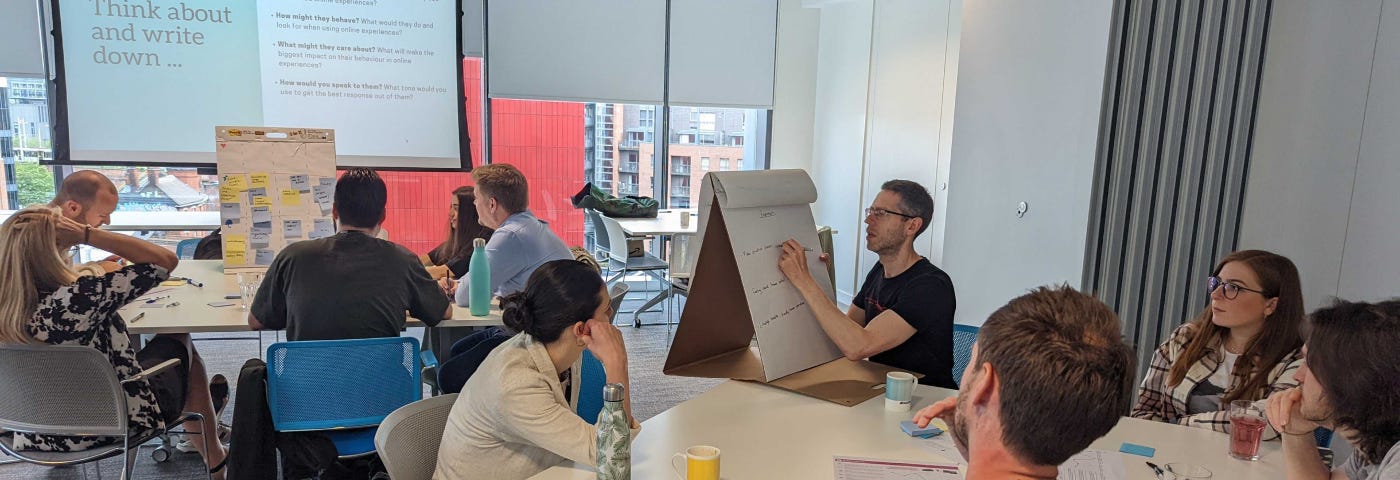 Two groups of four people having a conversation. Each group has one man writing ideas on a giant, white, sticky note.