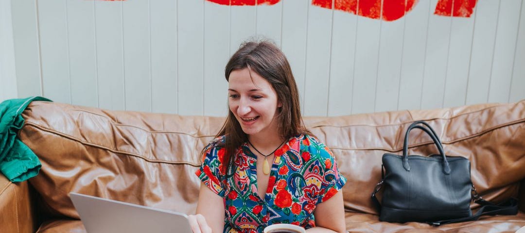 Picture of Rebecca Lockwood in a colourful dress, sitting on a sofa smiling as she looks at her open apple laptop screen. What NLP Training Can Do for You. NLP training, NLP coaching, and NLP techniques from, mother, an award-winning Master NLP Mindset Coach, Rebecca Lockwood.
