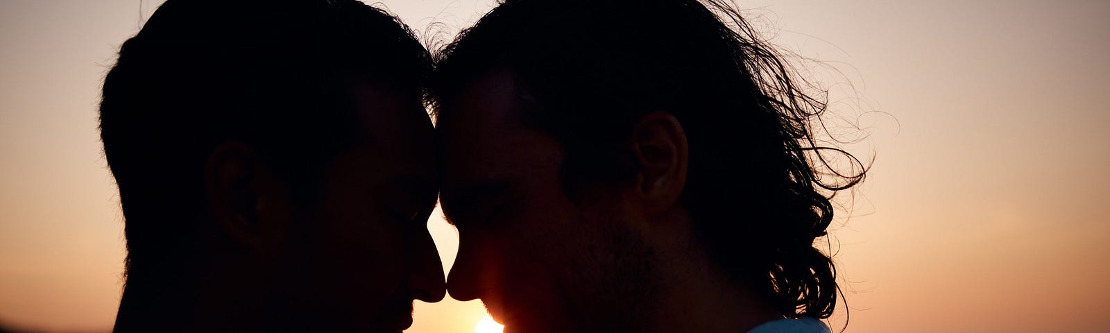 A silhouette of two men with their foreheads resting together and their noses touching with the sun setting in the background.