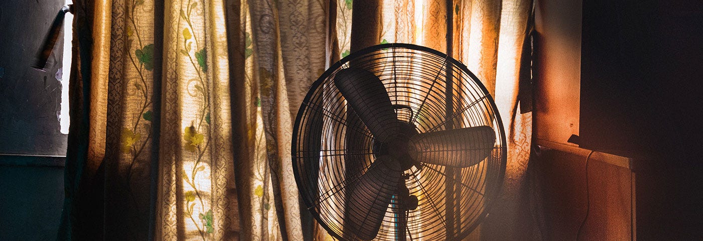 A household electric fan in front of curtains with a yellow and green floral design, lit by golden sunlight.