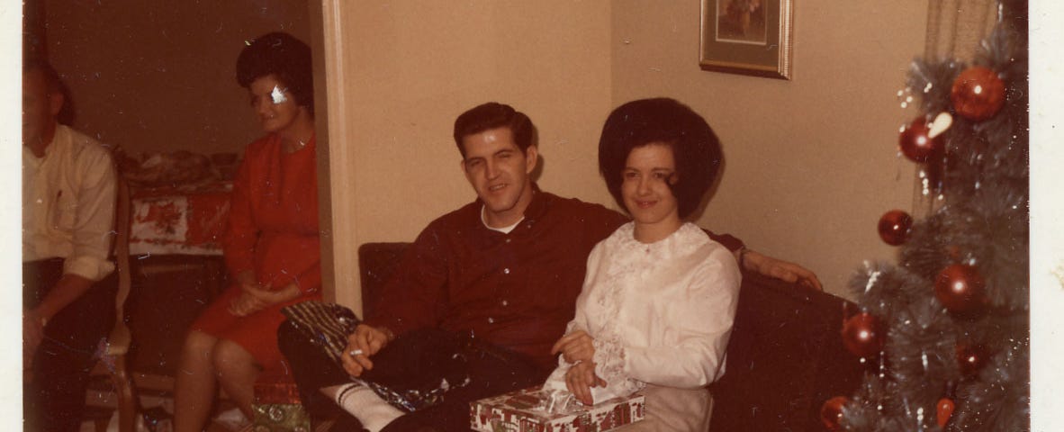 A 1960s-era couple sitting on a loveseat near a Christmas tree posing for the camera