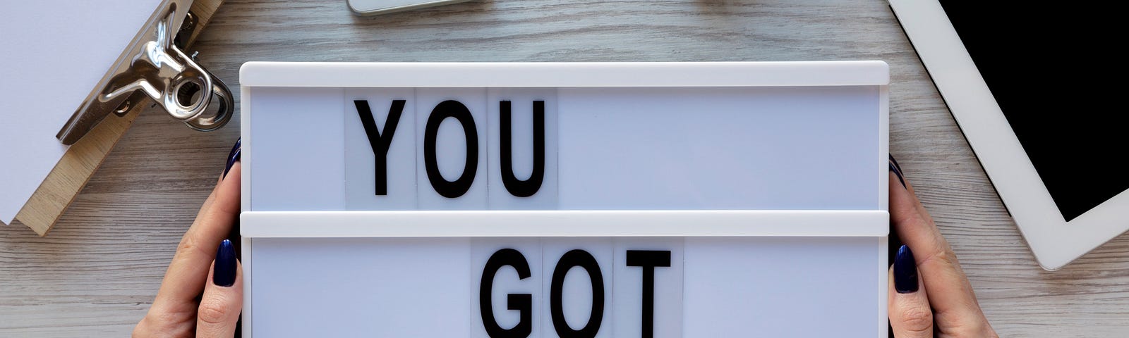 A person holds a sign that says, “You got this.” A notebook, iphone, and ipad are nearby on a gray desk.