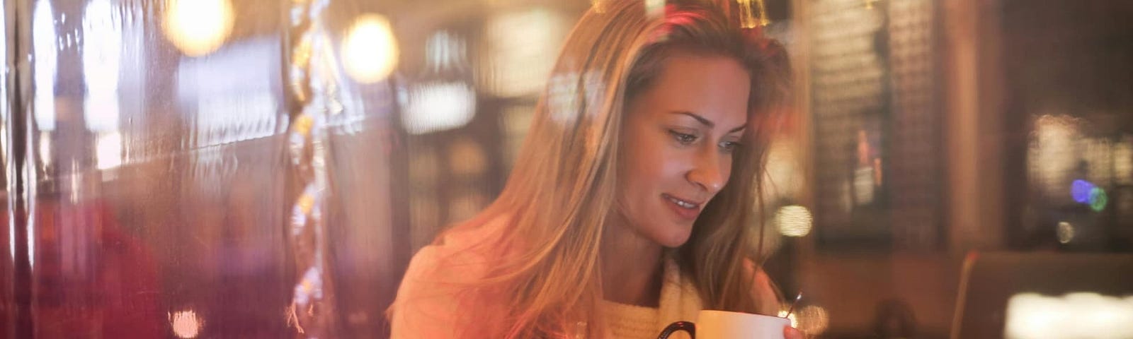 A girl with long hair sitting on a chair using her laptop with one hand while sipping a cup of coffee with another. The café has bright yellow bulbs and the image is taken behind a glass window.