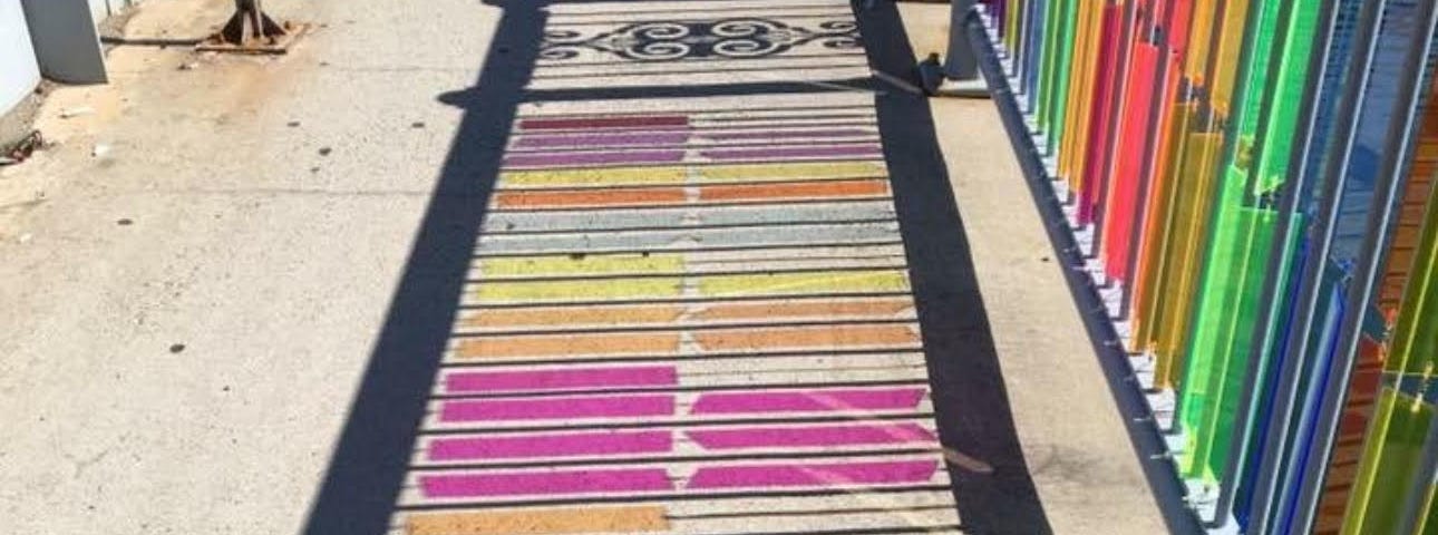 A photo of an art installation in Boston, on the Congress Street bridge, glass of various colors installation on the guardrail, reflecting colorfully on the concrete sidewalk.
