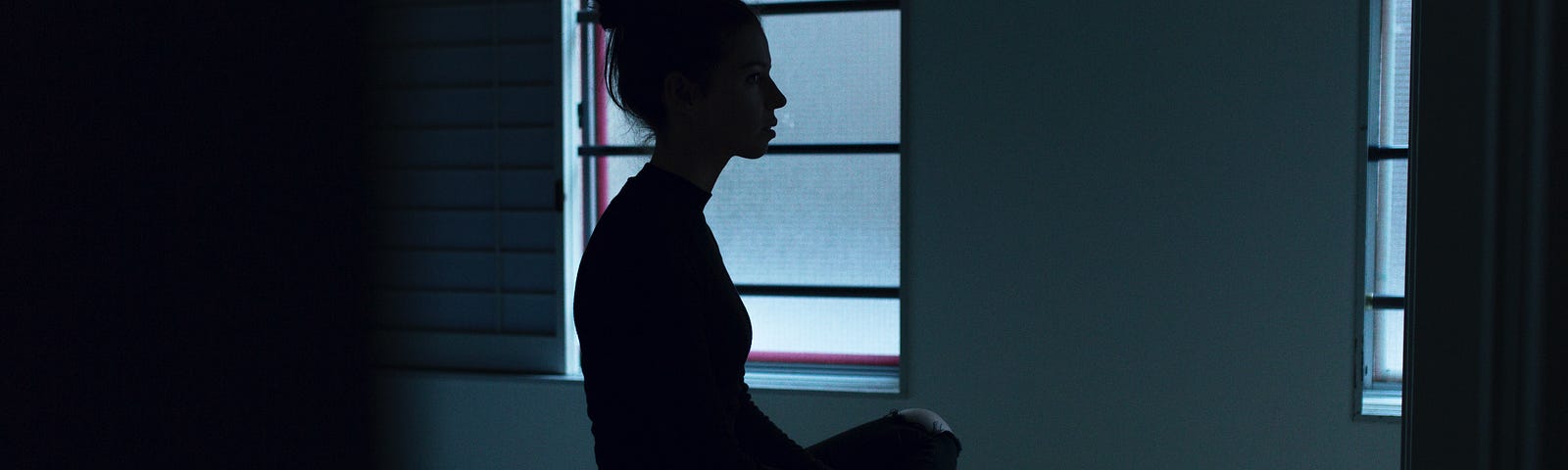 A silhouette of a woman with hair hair in a bun on her bed cross legged, looking at the wall at the end of her bed