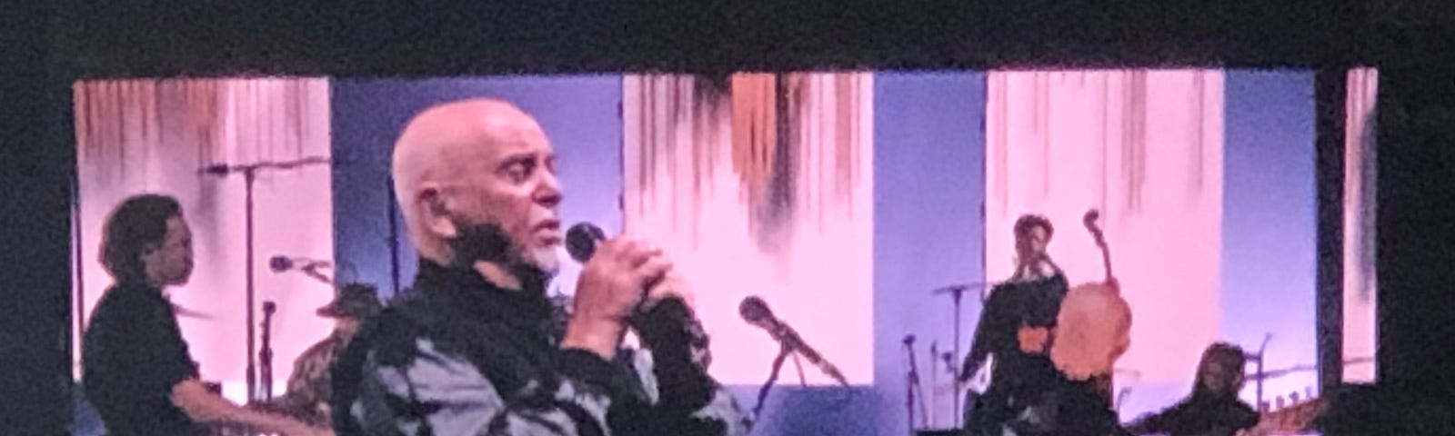 Peter Gabriel facing sideways, singing into a microphone with bandmates behind him