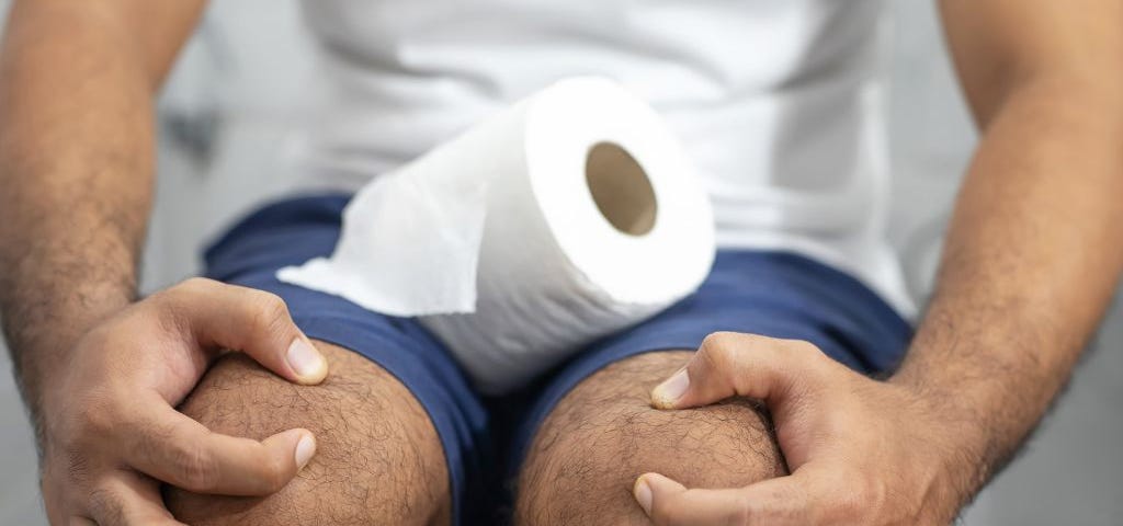 Man wearing blue shorts and white tshirt sitting on a toilet clutching his knees with a roll of toilet paper on his lap