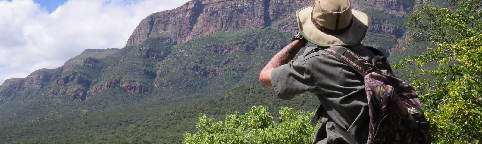 A Caucasian male wearing a trail guide uniform, sitting on a rock, looking through binoculars at a mountain range