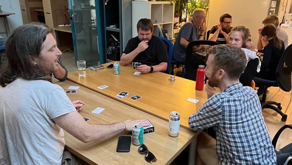 Four people sitting at a table playing a card based game, in the background another table of games players