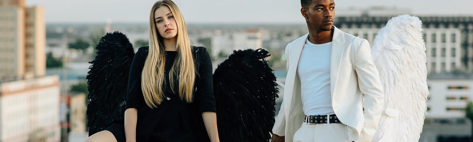 A white woman in a black angel costume and a Black man in a white angel costume hang out on a rooftop somewhere in a city. The woman looks straight at the camera. The man gazes to the side, into the distance.