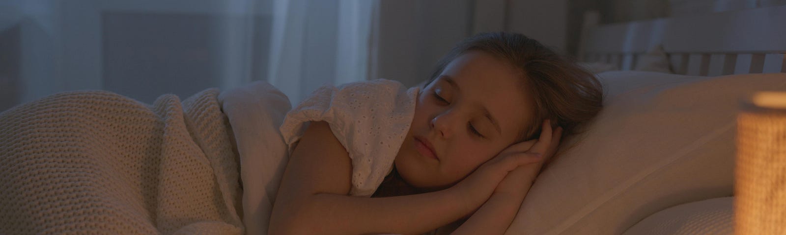 A young girl sleeps in her bed on her side. Her hands are clasped together under her head.