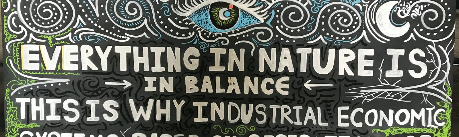 A large board painted black with swirly and branching patterns in white, green and blue, and the following words written in block capitals in white: Everything in nature is in balance. This is why industrial econcomic systems based on separation are destroying nature. Stories for life.