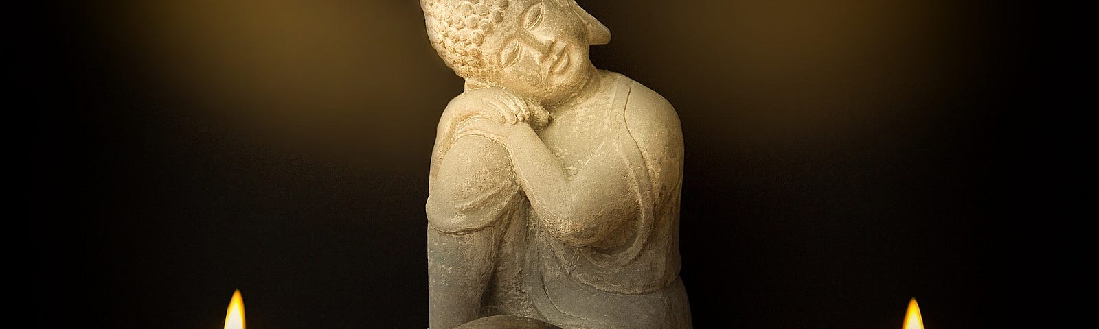 A small statue of Buddha is sitting with one knee close to his chest. His hands and ear are on that knee. In front of him are two rock candle holders with candles lit. Between the candles is a rock koan composed of four rocks.