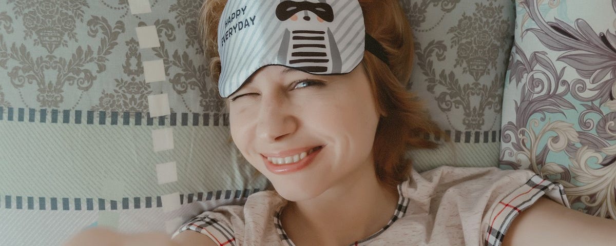 An attractive woman lying in bed, smiling and squinting at the camera. She is wearing pyjamas and has a sleep mask pushed up onto her forehead. The sleep mask shows a cartoon raccoon, and the words ‘Happy every day.’
