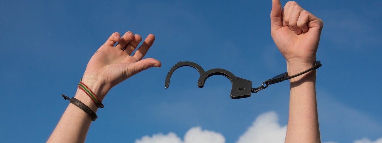 A photograph shows a person’s forearms raised up. Gray shirtsleeves are barely visible at the bottom of the image. The person has a woven or beaded multicolor bracelet and a dark colored leather bracelet on their right wrist. They have one locked handcuff on their left wrist. Attached is an open handcuff that swings in the air between their hands. A blue sky with puffy white clouds is shown in the background.