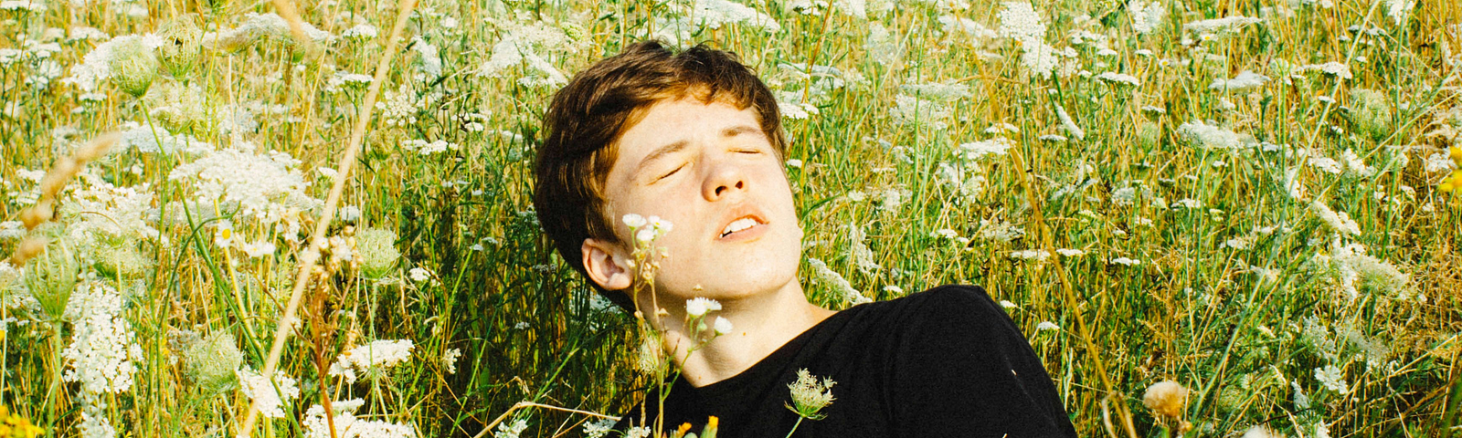 A young white man with short, slightly-ginger hair, wearing a black T-shirt and sitting and leaning back on his right arm in a field of tall green and amber grass and while flowers. It’s a sunny day, his eyes and closed and his face is turned towards the sun.