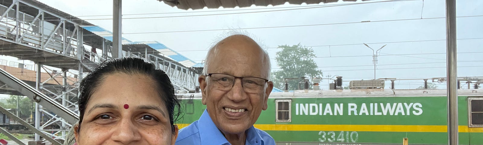 Selfie of the author, a woman in a green dress and grey scarf, with an older gentleman in a blue shirt, at a railway platform. There is an Indian Railways electric engine in the background. A railway platform overbridge is also visible in the background.