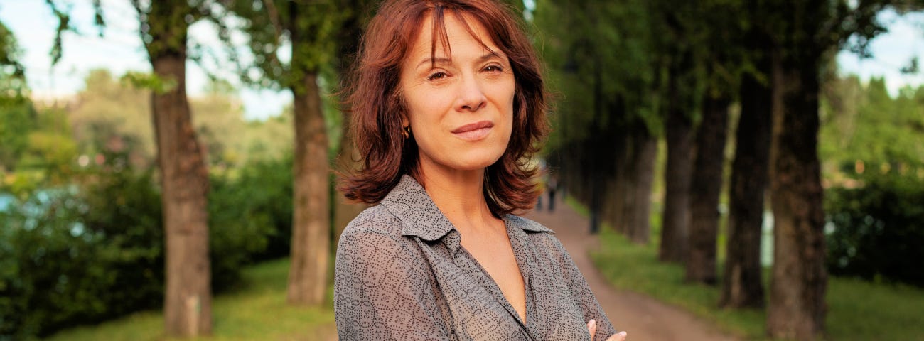 A woman with auburn hair standing on a dirt road with trees on either side looking annoyed.