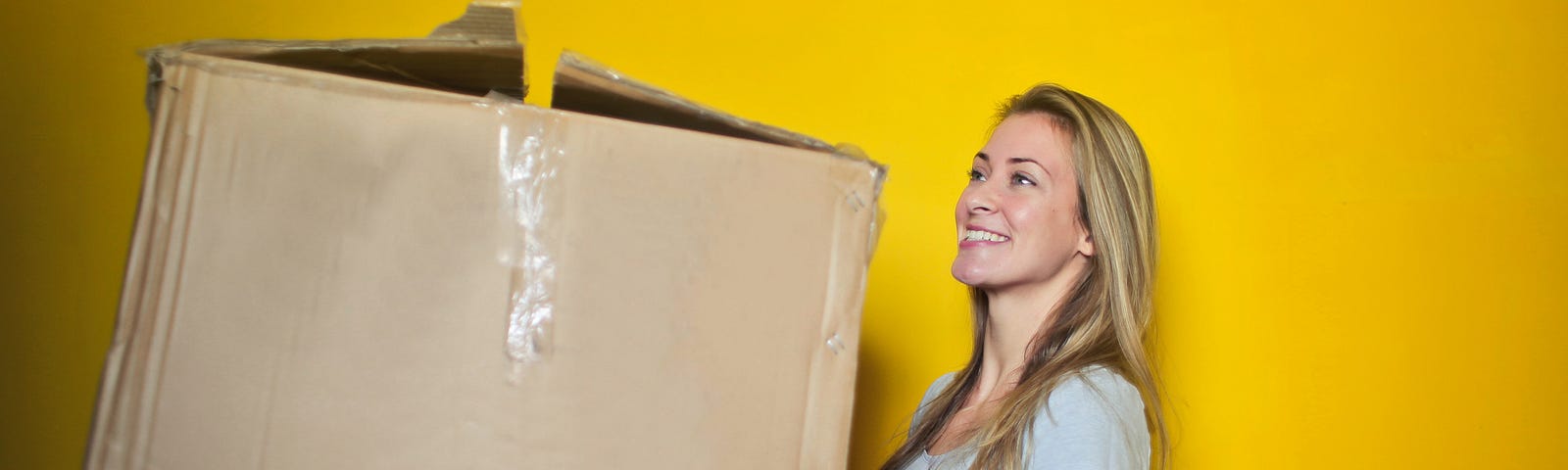 Woman In Grey Shirt Holding Brown Cardboard Box in front of a yellow wall.