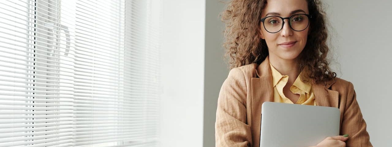 a woman in a beige blazer and glasses hugs a laptop to her body and smiles at the camera after comparing sex coach training programs