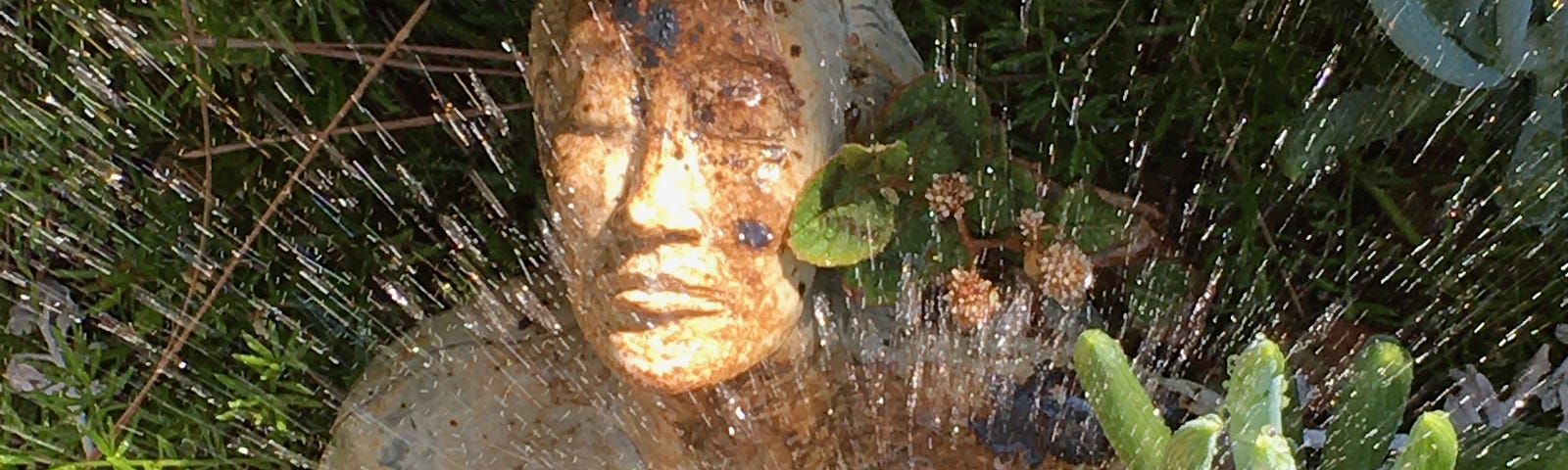 Sculpture of woma with hands reaching across chest together, eyes closed, sprinkler sprinkling water ovr her. She sits by cactus and lants.