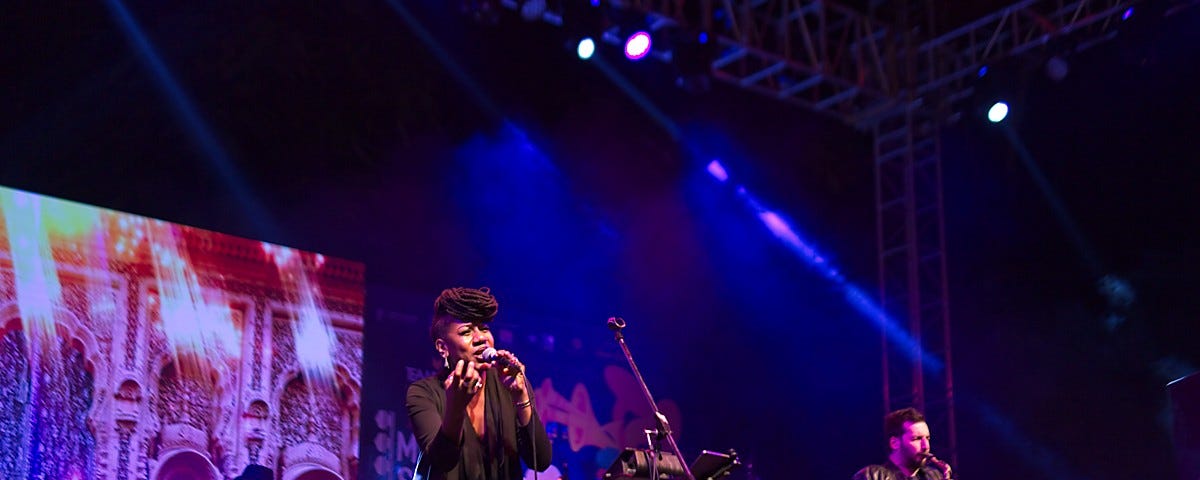 A large stage at night lit up with coloured lights, a bass player in the back ground, Black cis female center stage , microphone in hand, young saxophone player to her right.