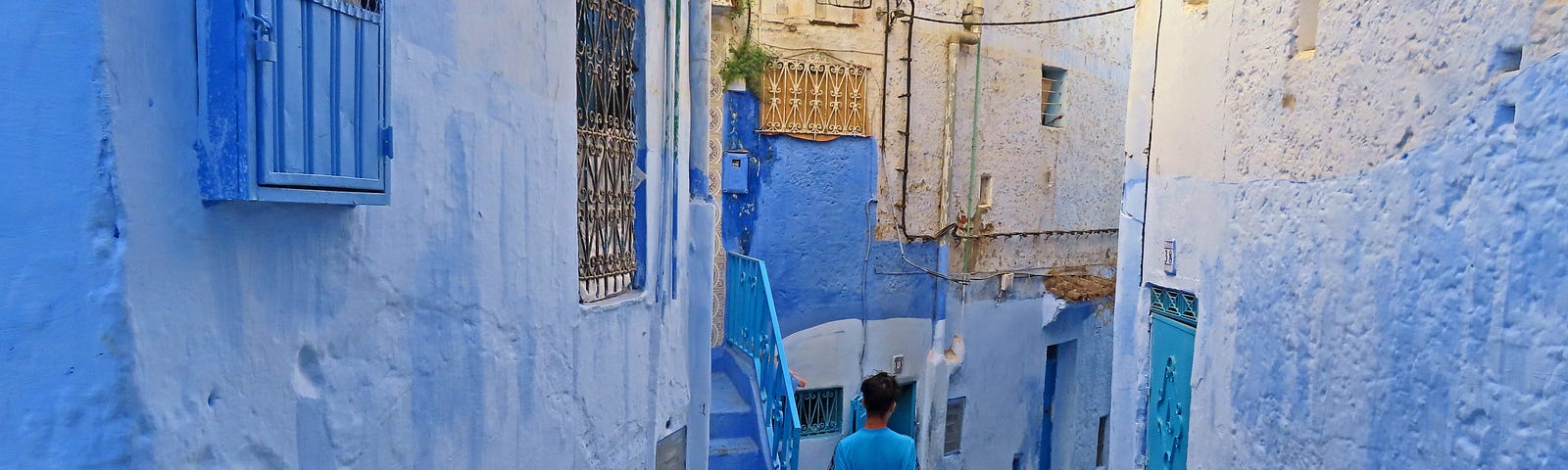 Man in a blue tshirt walks down a narrow street with blue, plastered buildings on either side. Blue doors, blue windows, blue stairs.