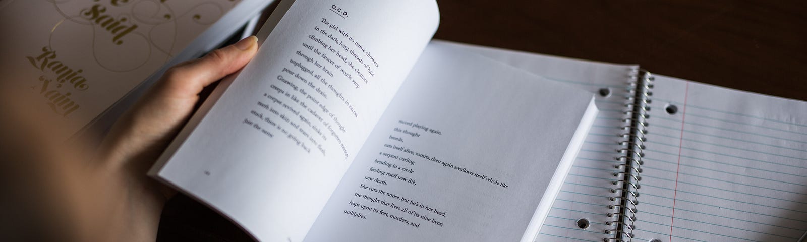 A young woman is pictured reading poetry from an open book, placed above empty notes from a large notebook.