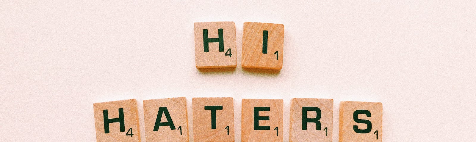 A picture of scrabble tiles that read “hi haters” on a light pink background.