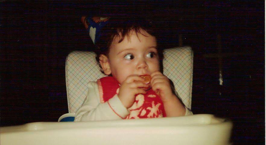 a baby sits in a retro-looking highchair with a party hat on and she’s biting into an orange segment with apparent trepidation