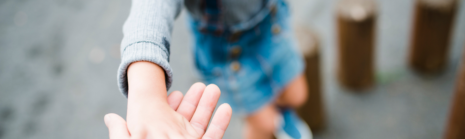 The image shows a close-up of one person holding another’s hand, helping or leading them along. The focus is on the clasped hands in the foreground, which is sharp and detailed against a blurred background. The child being led is only partly visible; we see them from the waist down, wearing denim shorts and blue shoes, standing on what appears to be a row of round wooden posts, possibly part of a playground or an outdoor balancing trail.