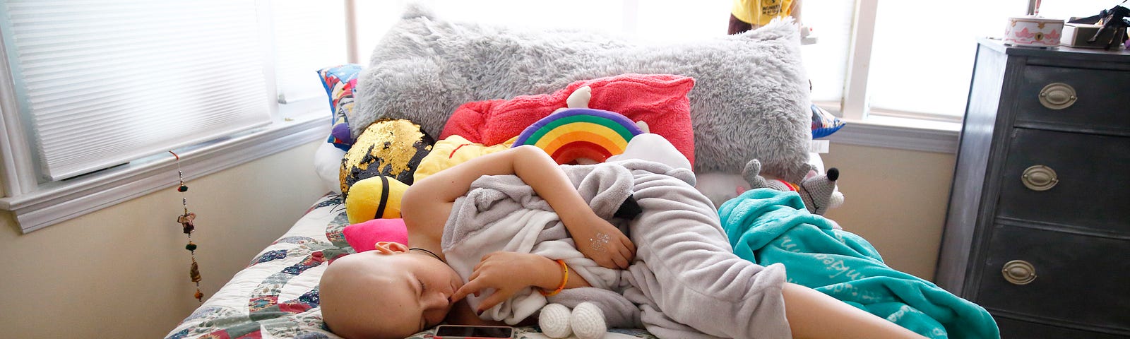 A young girl with cancer, lays on her bed looking a little nervous, surrounded by pillows and stuffed animals.