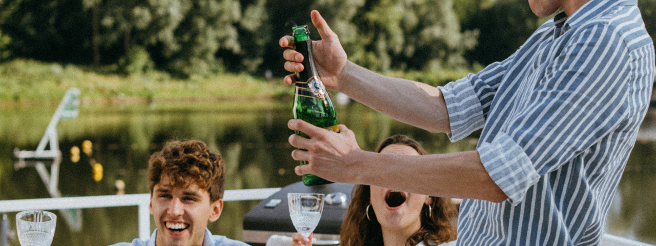 A man in a striped blue and white shirt pops open a bottle of champagne. Behind him a woman in a white shirt is holding a glass and looking up at him. Next to her is a man wearing a blue shirt and laughing. They are sat outside by a lake.