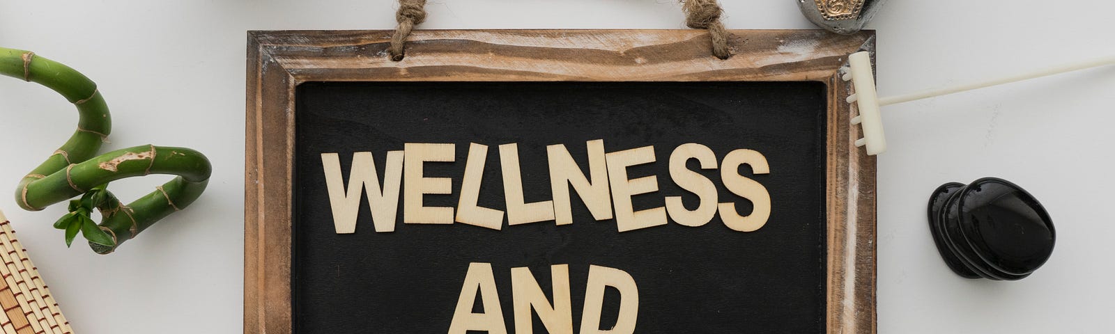 WELLNESS AND HEALTH written in all cap letters on a wood-framed board surrounded by curly bamboo, Buddha statues, succulent plants, balancing hot stones, rolled straw placemat, and incense in stone holder. Zen feel.