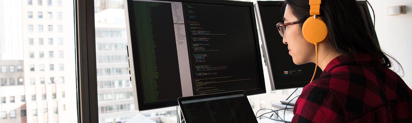 Woman wearing headphones writing code in front of a laptop and a large monitor