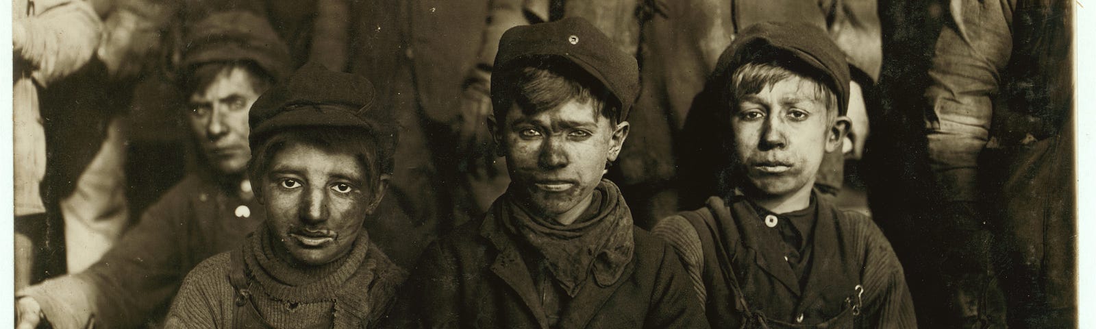 Lewis Hine b/w Library of Congress photo from 1911 showing child laborers, several young, seated  ‘breaker boys.’ The photo depicts small boys, 8–10 years old, in dirty clothing, faces sneared with coal dust, working in an anthracite coal mine in eastern Pennsylvania.